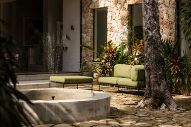 Sunlit stone courtyard patio with a modern green outdoor sofa and ottoman beside a bubbling round fountain, surrounded by tropical plants and a textured stone wall.