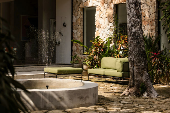 Sunlit stone courtyard patio with a modern green outdoor sofa and ottoman beside a bubbling round fountain, surrounded by tropical plants and a textured stone wall.