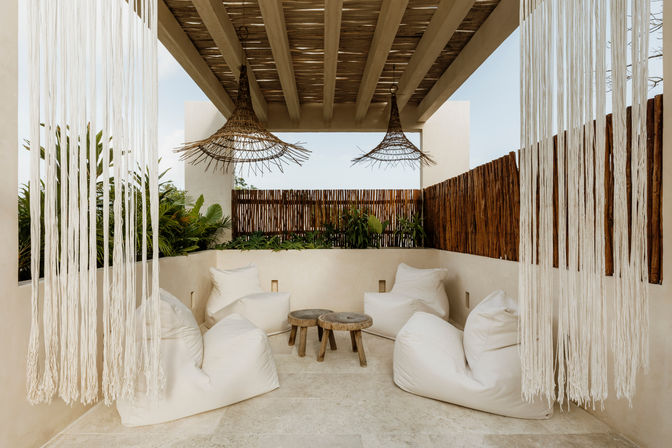 Boho outdoor patio under a wooden pergola with woven rattan pendant lights, macramé fringe curtains, cream beanbag chairs, low rustic wooden stools, tropical plants and a bamboo privacy fence.
