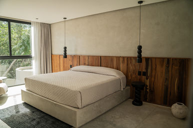 Sunlit minimalist bedroom with concrete platform bed, neutral quilted bedding, walnut wood headboard, two black pendant lights, small black side table and a large window framing green trees