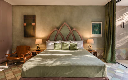 Sunlit green-themed bedroom with arched upholstered headboard, king bed with green pillows and throw, wooden nightstands with lamps, mid-century lounge chair and patterned tile floor