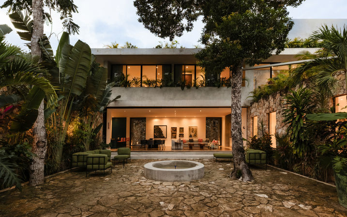 Modern two-story tropical villa courtyard at dusk with warm-lit open living room, central circular fountain, stone patio and green outdoor seating surrounded by lush palms and trees.