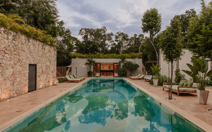 Inviting rectangular turquoise pool at a luxury tropical villa, flanked by wooden sun loungers, potted palms, stone walls and dense jungle under a cloudy sky.