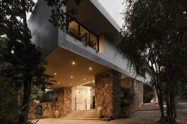 Cantilevered modern concrete home with stone-clad ground level and glass entry, nestled in lush trees at dusk with warm recessed lights illuminating steps and gravel driveway.