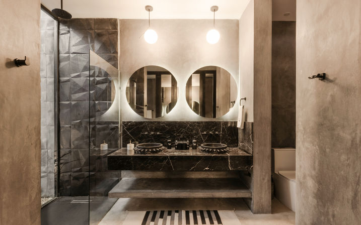 Sleek industrial-chic bathroom with black marble double vanity, two backlit round mirrors, pendant lights, concrete walls, black vessel sinks, and glass walk-in shower