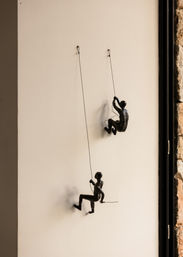 Playful black metal climber sculptures suspended by thin wires on a cream interior wall — modern indoor wall art beside a stone-framed window