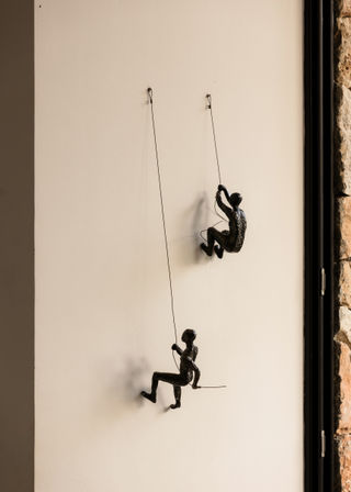 Playful black metal climber sculptures suspended by thin wires on a cream interior wall — modern indoor wall art beside a stone-framed window