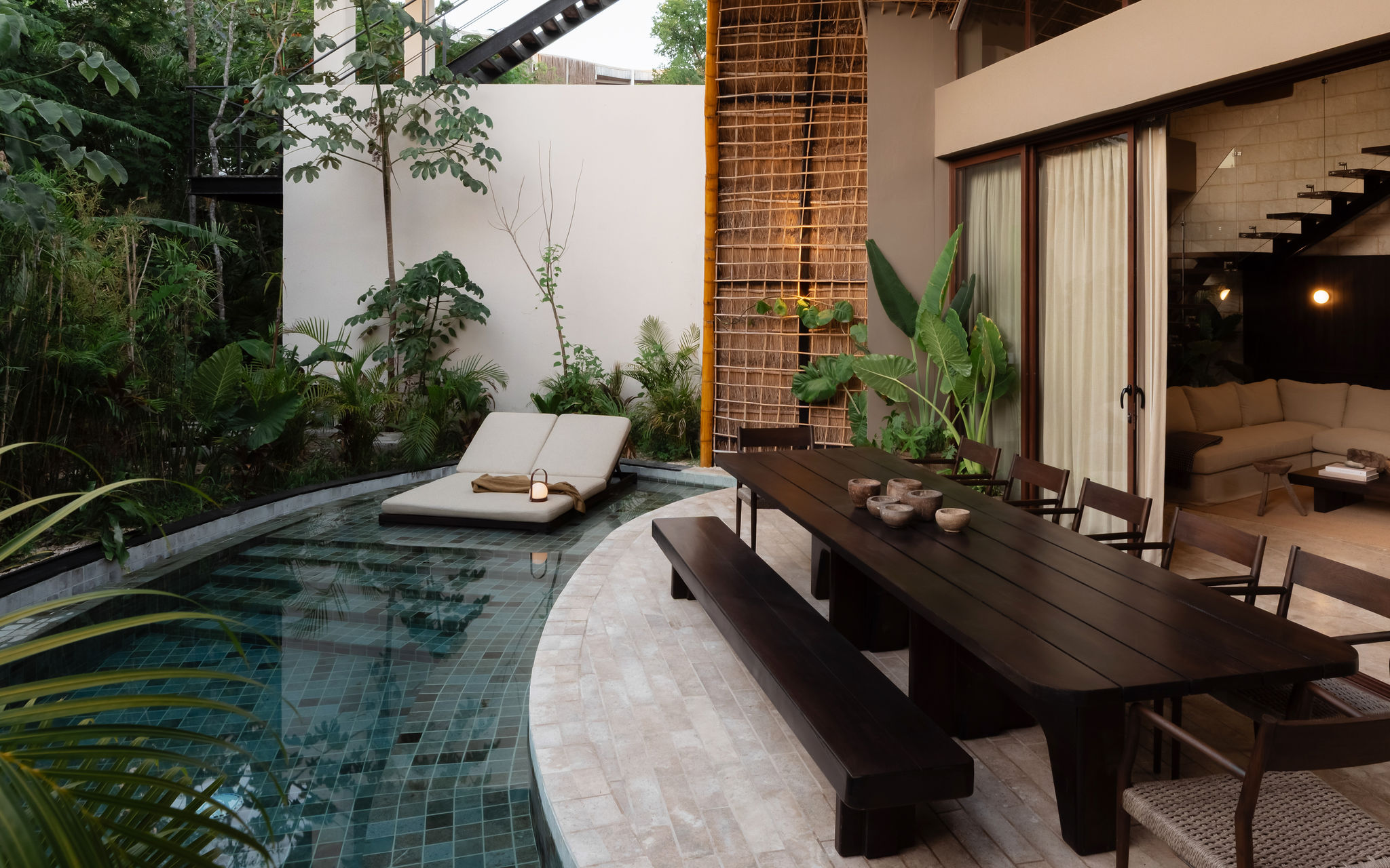 Curved tiled plunge pool at a tropical villa patio with a dark wood dining table and bench, cushioned sun loungers by the water, sliding glass doors to a cozy living room, and lush tropical plants with bamboo screening.