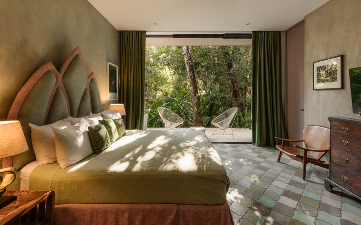 Sunlit green-themed bedroom with a large bed, textured headboard, patterned tile floor and mid-century chair, opening via sliding glass to a lush forest patio with two wire chairs — serene eco-retreat vibe.