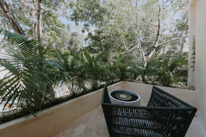 Cozy tropical balcony patio with woven wicker chair, circular tabletop fountain, planter box of palm plants and lush green trees beyond