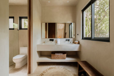 Sunlit modern minimalist bathroom with double vanity and integrated sinks, neutral plaster walls, wicker basket on shelf, walk-in shower, toilet, bench and large window overlooking trees.