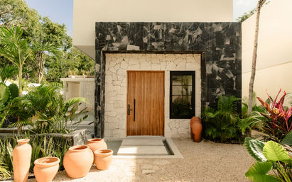 Sunlit modern tropical entrance with a vertical wooden door set in light stone and black marble frame, shallow reflecting pool, clustered terracotta pots, and lush palm and tropical foliage.