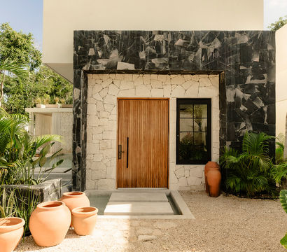 Sunlit modern tropical entrance with a vertical wooden door set in light stone and black marble frame, shallow reflecting pool, clustered terracotta pots, and lush palm and tropical foliage.