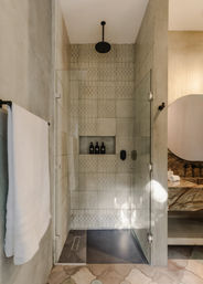 Spa-like modern walk-in shower with frameless glass door, black rain showerhead, patterned neutral tiles and recessed niche holding three black bottles, beside a stone vanity and white towel.