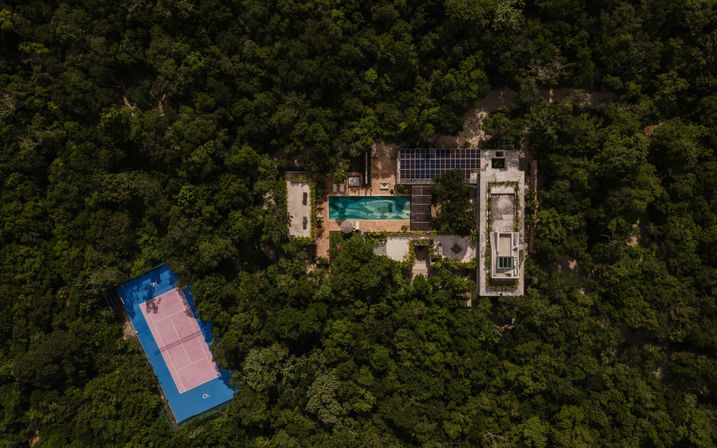 Aerial view of a hidden jungle oasis: modern estate with a long turquoise swimming pool, rooftop solar panels, and a blue-and-pink tennis court surrounded by dense tropical forest.