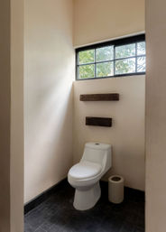 Minimalist small bathroom with white one-piece toilet, black tile floor, beige plaster walls, two rustic wooden floating shelves and a high black-framed window letting in natural light and views of greenery.