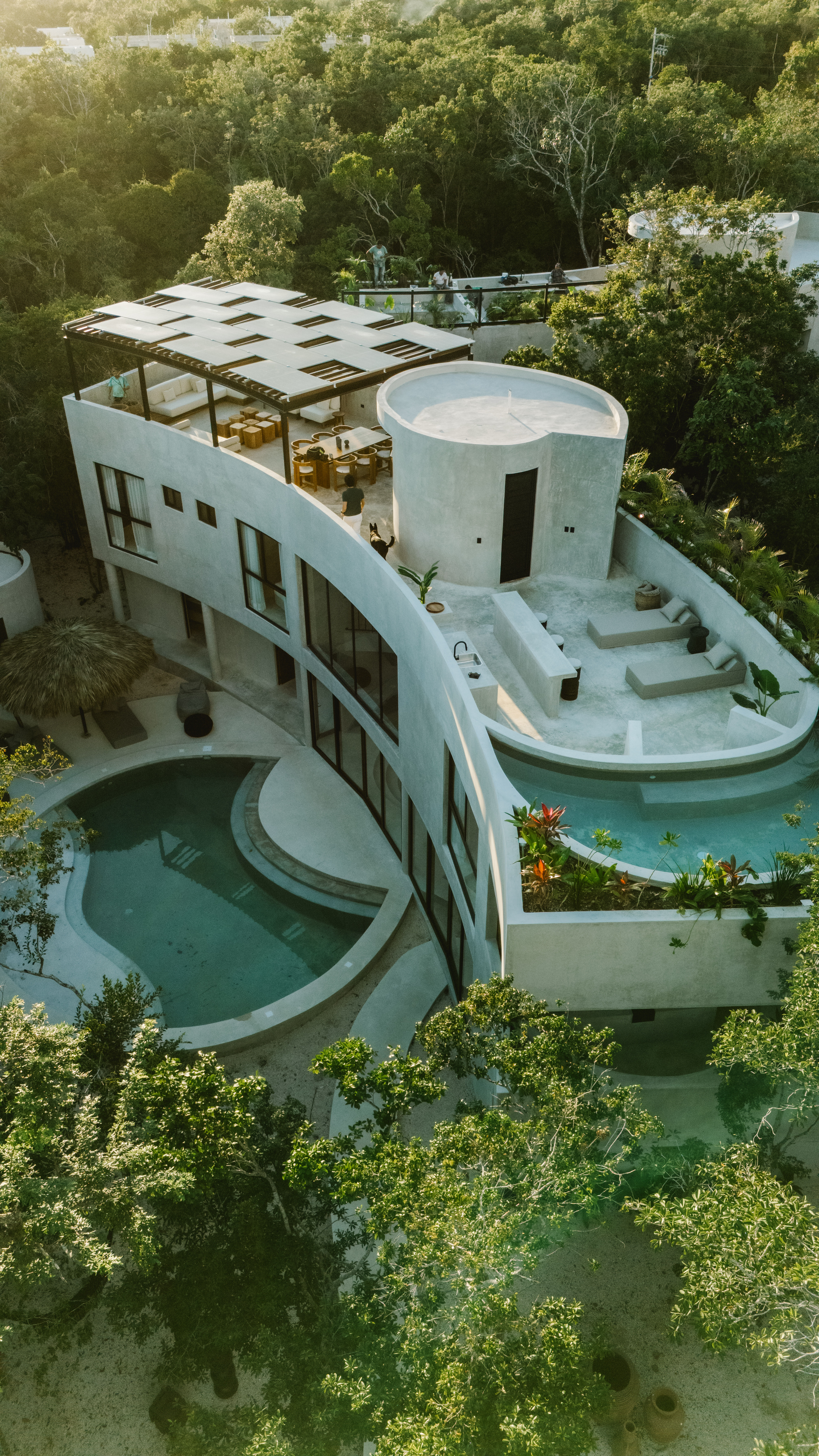 Aerial view of a sunlit modern curved concrete villa on a lush tropical hillside, featuring a rooftop pergola dining area, terraced lounge and two curved swimming pools surrounded by dense green forest.