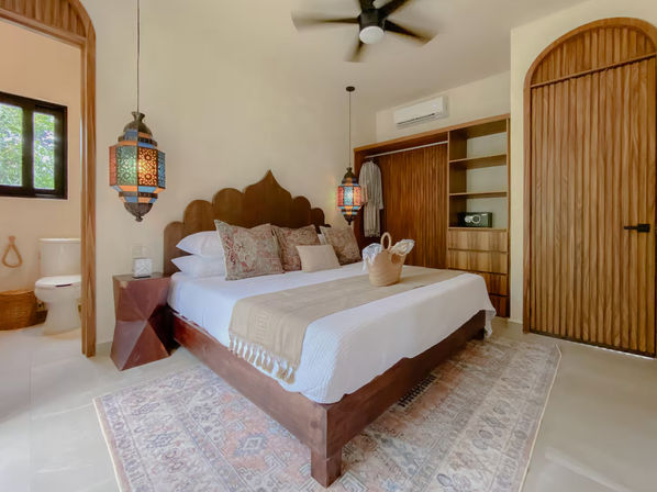 Sunlit boho bedroom with scalloped wooden headboard, king bed in white linens and patterned pillows, colorful Moroccan pendant lamps, wooden wardrobe and arched door, ceiling fan, patterned area rug and visible ensuite bathroom.