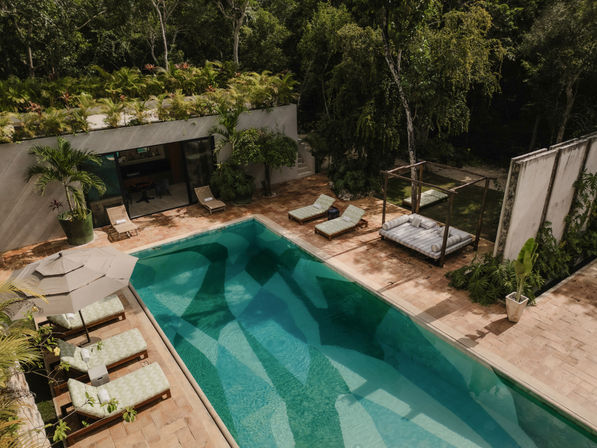 Aerial view of a turquoise rectangular swimming pool at a modern villa terrace with cushioned sun loungers, shaded umbrella, canopy daybed, stone tile patio and lush tropical greenery