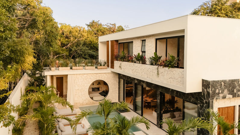 Sunlit modern two-story tropical villa with private plunge pool, outdoor lounge and palm trees; white stucco and stone facade with a round window, balcony planters and floor-to-ceiling glass doors surrounded by lush greenery.