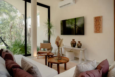 Sunlit modern tropical living room with floor-to-ceiling glass doors and garden view, white sofa with mauve cushions, round wooden coffee tables, woven chair, tall indoor plant, wall-mounted TV and ceramic vases.