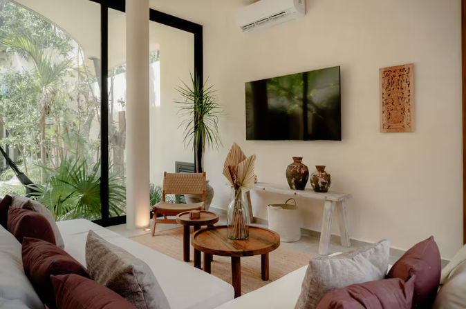 Sunlit modern tropical living room with floor-to-ceiling glass doors and garden view, white sofa with mauve cushions, round wooden coffee tables, woven chair, tall indoor plant, wall-mounted TV and ceramic vases.