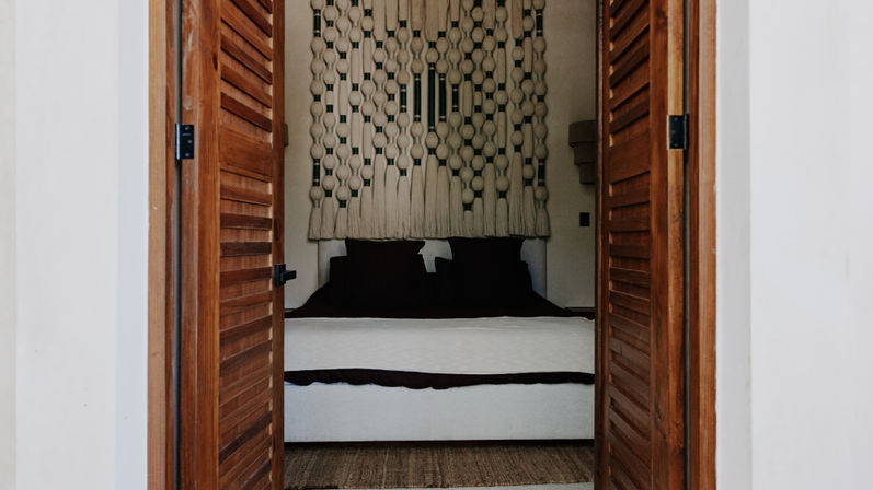 View through open wooden louvered doors into a cozy boho bedroom with white linens, dark pillows, woven rug and a large macramรฉ wall hanging.