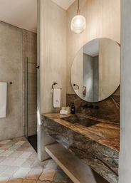 Modern spa-like bathroom with floating brown marble vanity and integrated sink, large round mirror, ribbed glass pendant light, matte black fixtures, textured plaster walls, pastel geometric floor tiles and glass walk-in shower