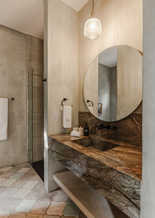 Modern spa-like bathroom with floating brown marble vanity and integrated sink, large round mirror, ribbed glass pendant light, matte black fixtures, textured plaster walls, pastel geometric floor tiles and glass walk-in shower
