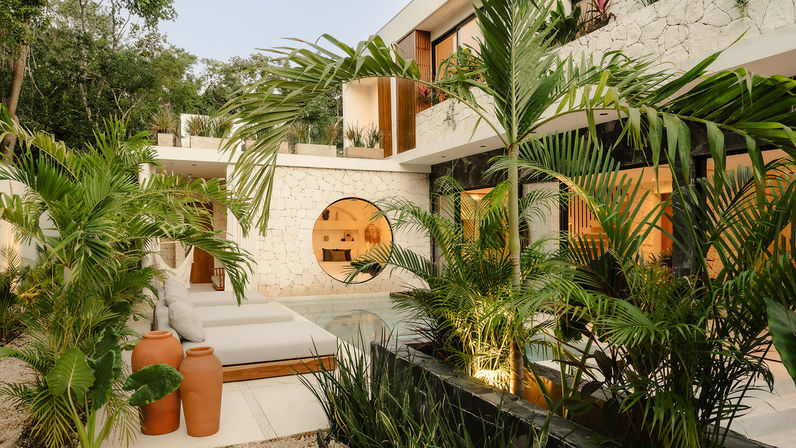 Sunlit tropical modern courtyard with a plunge pool, cushioned daybeds, terracotta pots, lush palm plants, and a stone wall featuring a round window with warm interior lighting.