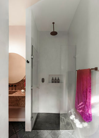Modern minimalist bathroom with glass walk-in shower and ceiling rainfall showerhead, built-in shampoo niche, bright magenta towel on wall, round mirror and stone vanity visible through doorway, pale plaster walls and patterned floor tiles.