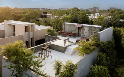 Sunset-lit rooftop terrace of a modern concrete villa with a small plunge pool, wooden bar stools, outdoor sofa and lush tropical greenery overlooking a residential neighborhood.