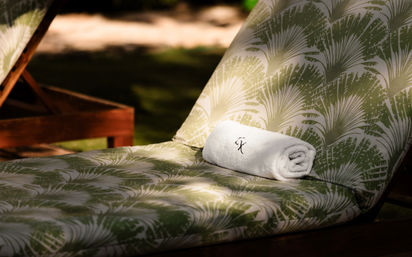 Sun-dappled wooden lounge chair with green palm-print cushion and a neatly rolled white embroidered towel, tropical poolside vibe.