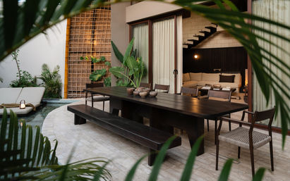 Modern tropical poolside patio with a dark wooden outdoor dining table, long bench and woven chairs, potted greenery, lounge chairs by a small pool, and sliding glass doors opening to a cozy living room and staircase.