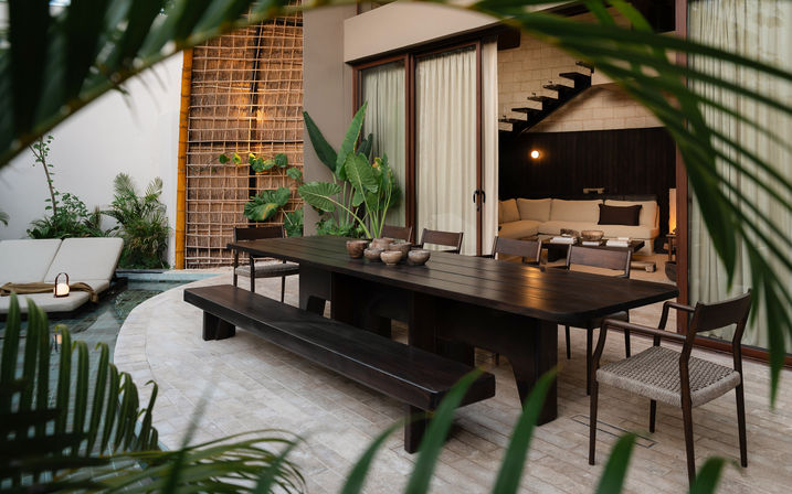 Modern tropical poolside patio with a dark wooden outdoor dining table, long bench and woven chairs, potted greenery, lounge chairs by a small pool, and sliding glass doors opening to a cozy living room and staircase.