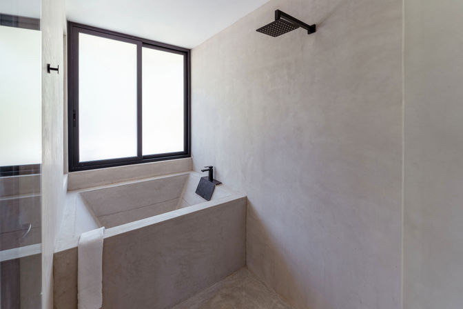 Modern minimalist bathroom with a concrete soaking tub, frosted window, matte-black waterfall faucet and square rain showerhead