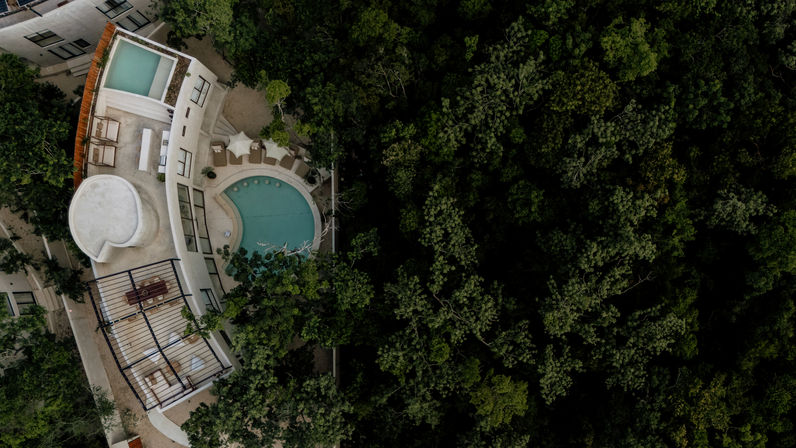 Aerial view of a curved modern villa with a circular swimming pool and rooftop terrace nestled at the edge of a dense green forest canopy.