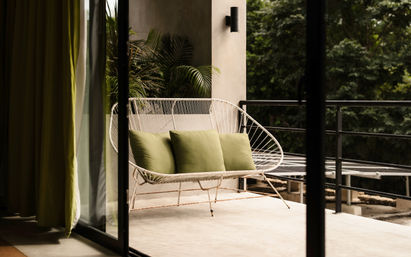 Sunlit modern balcony with a white woven outdoor loveseat topped with three olive-green cushions, sliding glass door, potted palms and leafy tree view.