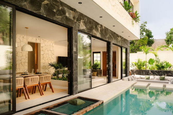 Modern tropical villa patio with large sliding glass doors revealing a stone-walled dining room and living area, turquoise rectangular pool with sunken lounge and netted pool hammock, and lush palm plants.