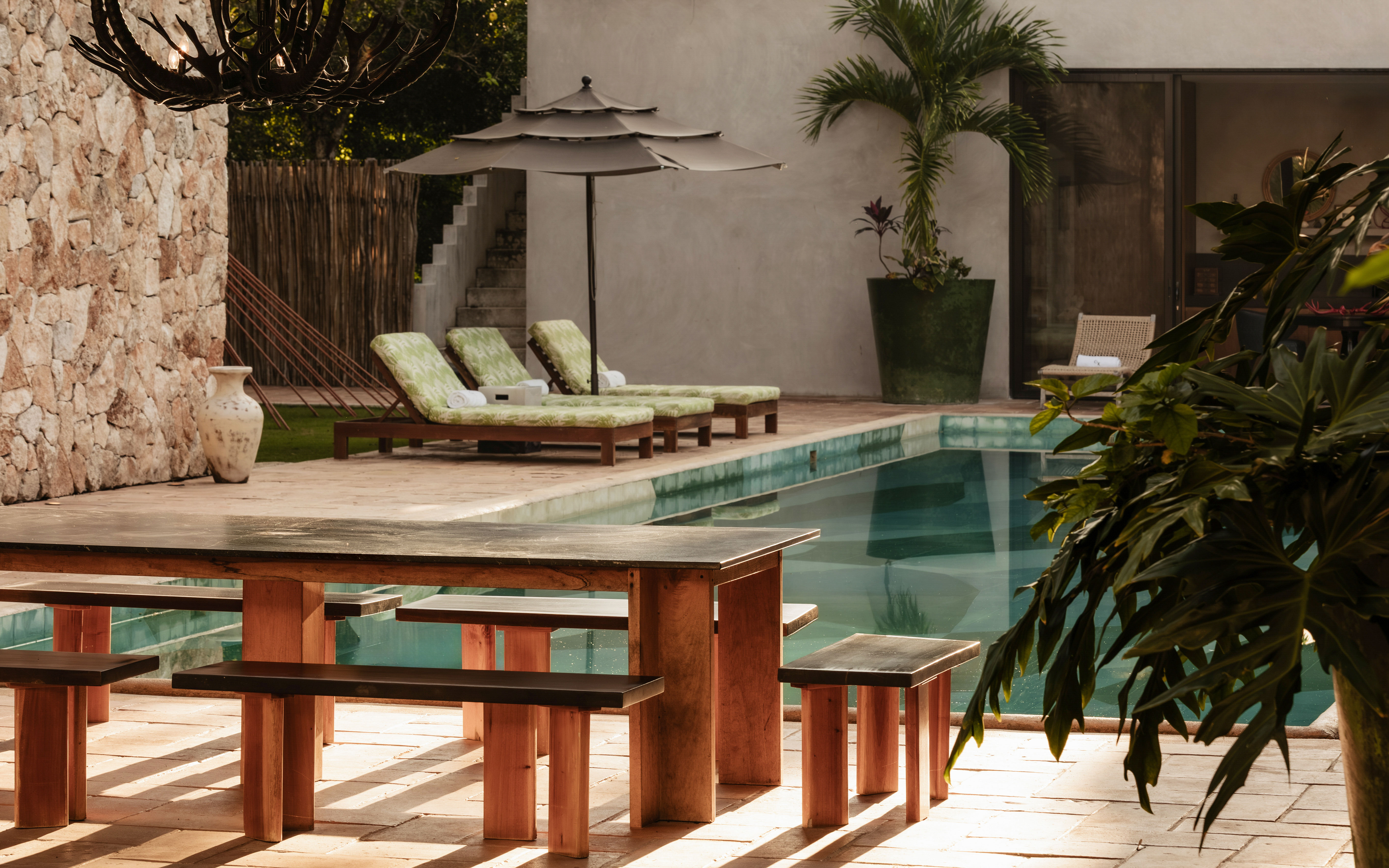 Sun-soaked tropical patio with a rectangular swimming pool, wooden picnic table and benches, green cushioned lounge chairs under an umbrella, stone wall and potted palm