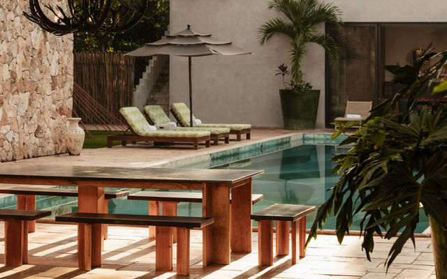 Sun-soaked tropical patio with a rectangular swimming pool, wooden picnic table and benches, green cushioned lounge chairs under an umbrella, stone wall and potted palm