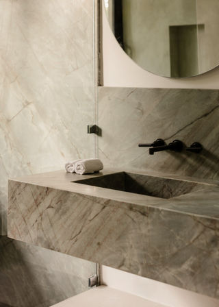 Spa-style marble bathroom sink with integrated rectangular basin, matte-black wall faucet, round mirror and rolled white towels