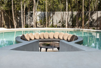 Resort-style sunken circular lounge with round beige cushions and a low coffee table inside a turquoise outdoor pool, framed by concrete decking and tall tropical trees.