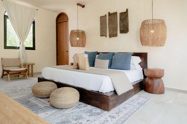 Sunlit boho bedroom with low dark-wood platform bed, white linens and blue pillows, woven pendant lights, round wicker poufs and patterned rug — airy coastal-inspired retreat.