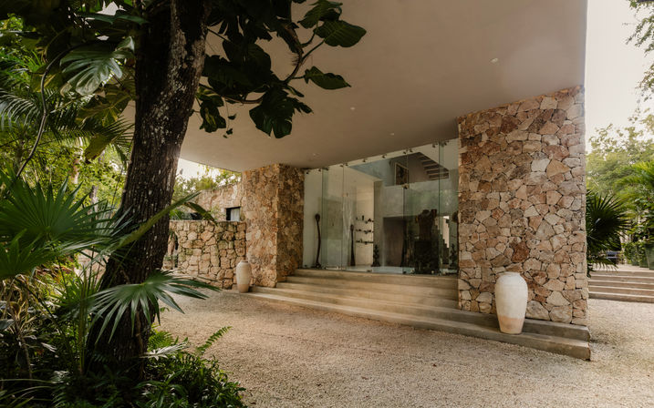 Modern tropical stone villa entrance with glass facade, concrete steps, terracotta urns, gravel courtyard and lush jungle foliage