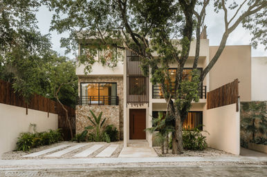 Two-story contemporary tropical home exterior with stone cladding, wood slatted screen, large windows, gravel driveway with concrete strips and lush landscaping under mature trees.