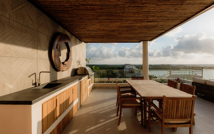 Covered ocean-view rooftop terrace with built-in outdoor kitchen and sink, long wooden dining table and chairs, glass railing and waterfront sunset view.