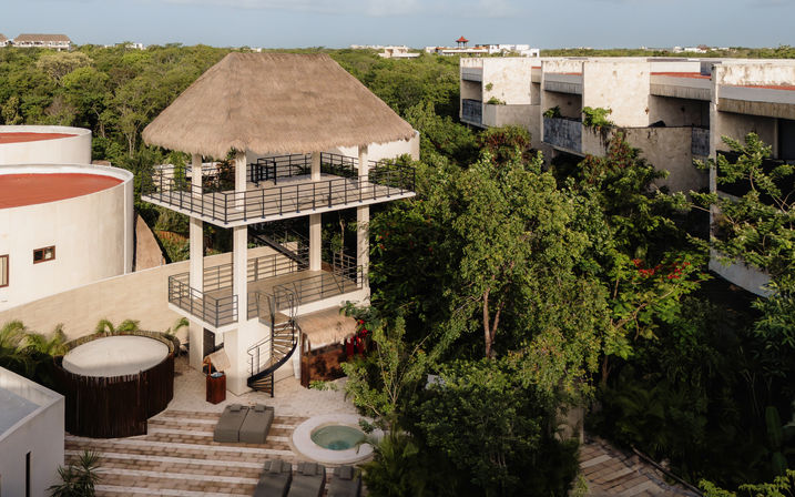 Aerial view of a three-level thatched-roof palapa tower with a spiral staircase overlooking a sunlit resort courtyard with a small circular plunge pool, lounge chairs and dense tropical greenery, with modern concrete buildings in the background.