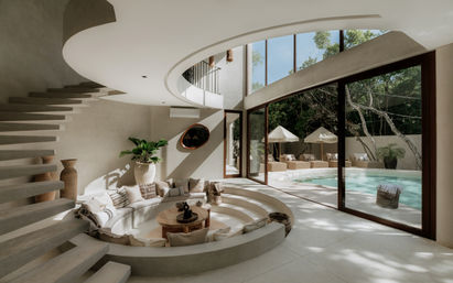 Sunlit modern villa interior with a circular sunken seating area framed by neutral cushions, a floating curved staircase, and floor-to-ceiling glass doors opening to a private poolside patio with lounge beds, umbrellas, and lush greenery.