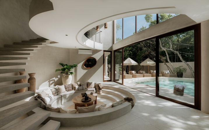 Sunlit modern villa interior with a circular sunken seating area framed by neutral cushions, a floating curved staircase, and floor-to-ceiling glass doors opening to a private poolside patio with lounge beds, umbrellas, and lush greenery.